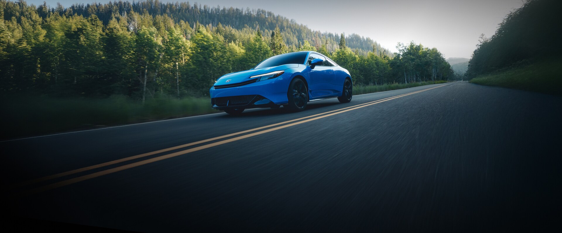 2026 Honda Prelude in Boost Blue Pearl driving trough a road with trees in the background.