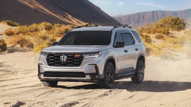 2026 Honda Pilot in Solar Silver Metallic front driving through a sandy desert landscape (Full-Size SUV).