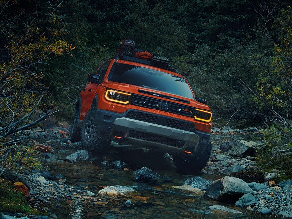 2027 Honda Passport in Sunset Orange profile driving through a rocky stream in a forest (Mid-Size SUV).