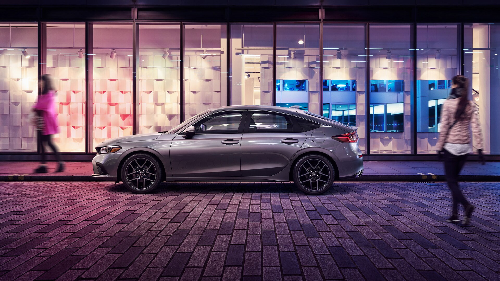 Driver-side profile view of the 2022 Honda Civic Sport Touring Hatchback in Lunar Silver Metallic, parked on a city street as the driver approaches.