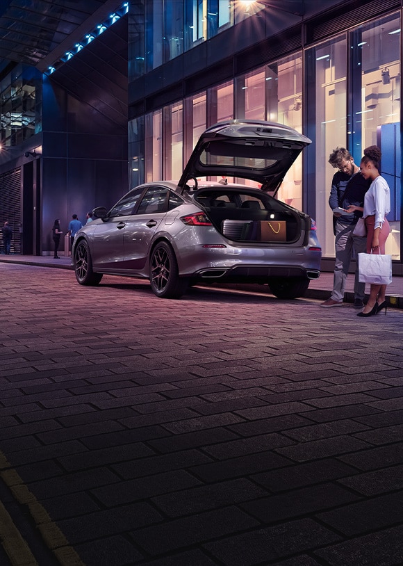 Rear driver-side view of the 2022 Honda Civic Sport Touring Hatchback in Lunar Silver Metallic, parked outside a store with the hatch open and a man and woman loading cargo.