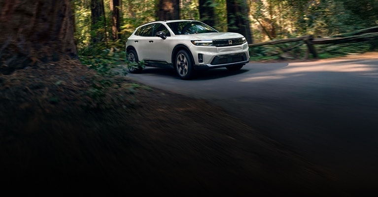 2026 Honda Prologue Elite in Frostline White Pearl front driving through a dense forest on a paved road (All-Electric SUV).