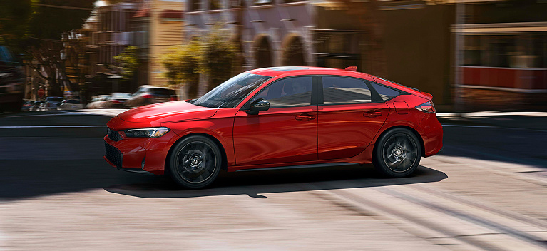 2026 Honda Civic Hatchback Sport Hybrid in Rallye Red profile driving in a blurred city street (Compact Hatchback).