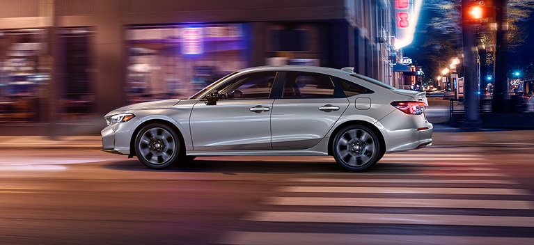 2026 Honda Civic Sedan Sport Touring Hybrid in Solar Silver Metallic profile driving on a city street (Compact Sedan). 2026 Honda Civic Sedan Sport Touring Hybrid in Solar Silver Metallic profile driving on a city street (Compact Sedan).