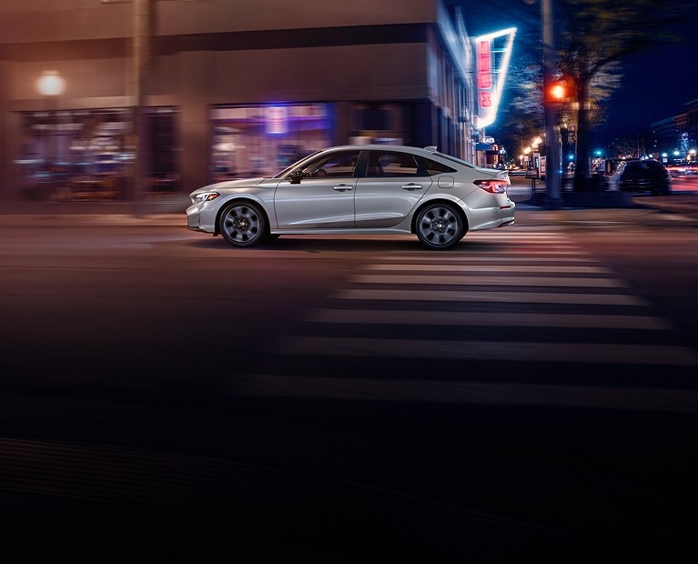 2025 Honda Civic Sedan Sport Touring Hybrid in Solar Silver Metallic driving on urban street at night - Opens a dialog