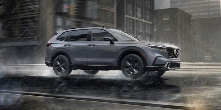 2027 Honda CR-V Sport Touring Hybrid in Urban Gray Pearl profile driving on a rainy city street (Compact Crossover SUV).
