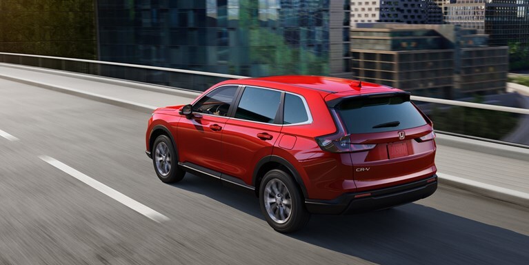 2027 Honda CR-V EX in Radiant Red Metallic back driving on a city bridge through the downtown skyline (Compact Crossover SUV).