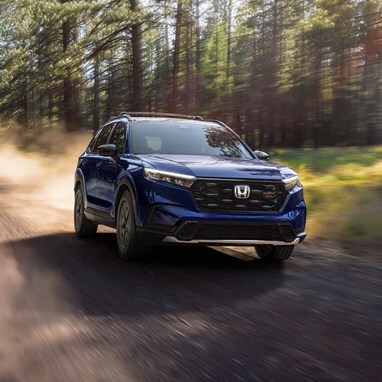 2027 Honda CR-V TrailSport Hybrid in Canyon River Blue Metallic front driving through a sunlit forest on a dirt road (Compact Crossover SUV).