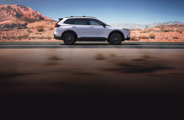 2027 Honda CR-V Sport Touring Hybrid in Platinum White Pearl profile driving on a scenic desert road (Compact Crossover SUV).