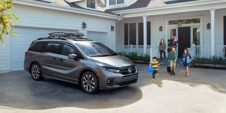 2027 Honda Odyssey Elite in Modern Steel Metallic front parked in a residential driveway with a family (Minivan).