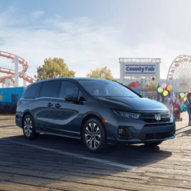 2027 Honda Odyssey Elite in Smoke Blue Pearl front parked on a wooden boardwalk at a county fair (Minivan).