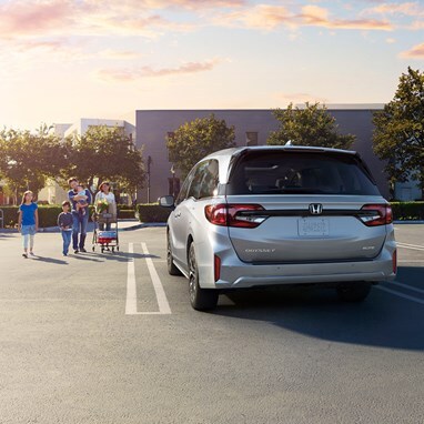 2027 Honda Odyssey Elite family van in Solar Silver Metallic back parked in a parking lot with a family walking nearby (Minivan).