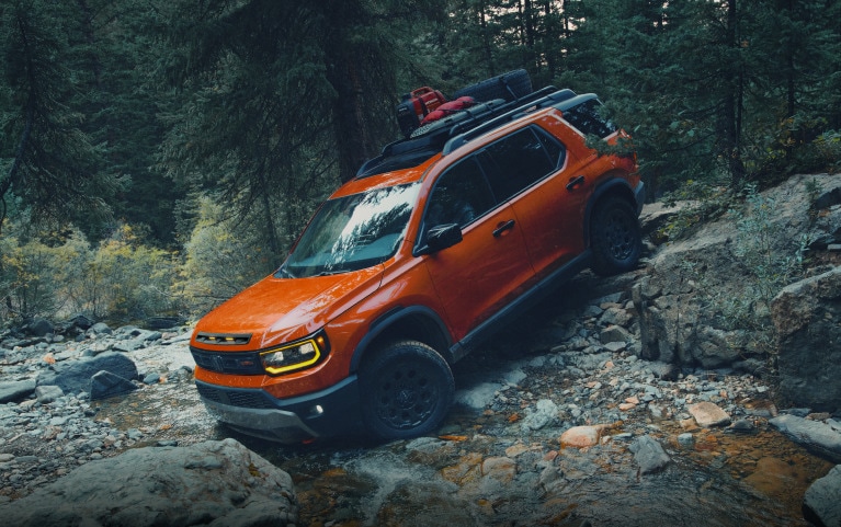 2027 Honda Passport TrailSport Elite in Sunset Orange profile on a rocky trail in a dense forest (Mid-Size SUV).