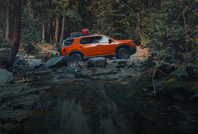 2027 Honda Passport TrailSport Elite in Sunset Orange profile crossing a rocky forest river with gear (Mid-Size SUV). 2027 Honda Passport TrailSport Elite in Sunset Orange profile crossing a rocky forest river with gear (Mid-Size SUV).