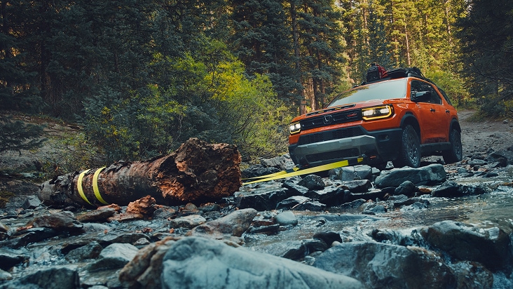 2027 Honda Passport TrailSport Elite in Sunset Orange front rocky riverbed towing a large log in the forest (Mid-Size SUV). 2027 Honda Passport TrailSport Elite in Sunset Orange front rocky riverbed towing a large log in the forest (Mid-Size SUV).