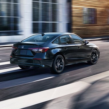 2027 Honda Civic Sedan Sport Hybrid in Crystal Black Pearl rear three-quarter view driving on a city street (Compact Sedan)